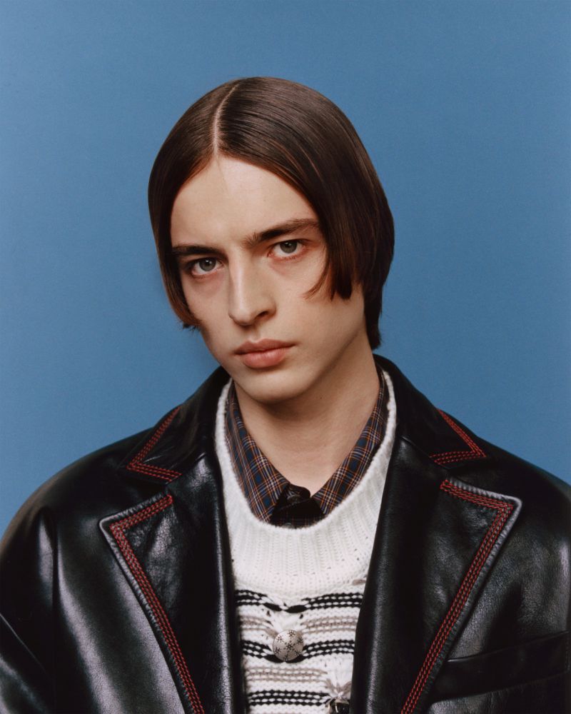 Portrait of a young adult male with dark brown hair, wearing a black leather jacket over a sweater and collared shirt, against a blue background.