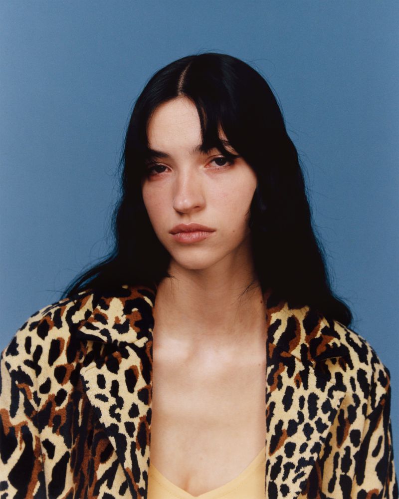Portrait of a young woman with dark hair, wearing an animal print coat against a blue background, studio lighting, fashion photography.