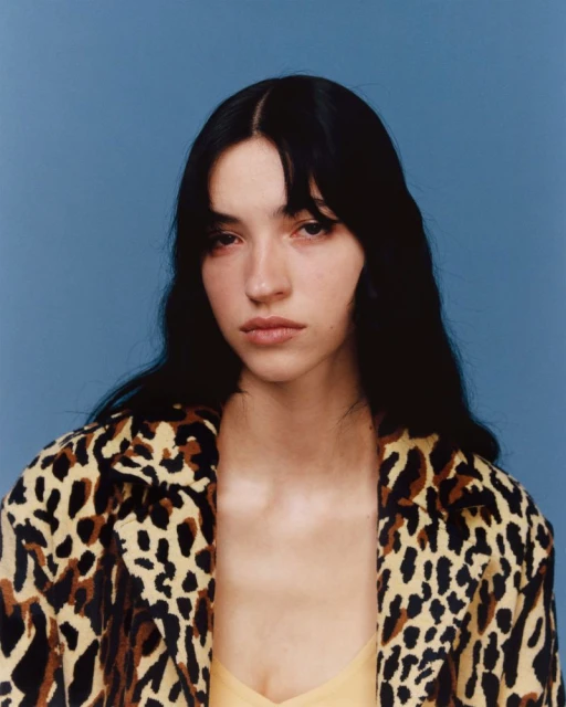 Portrait of a young woman with dark hair, wearing an animal print coat against a blue background, studio lighting, fashion photography.