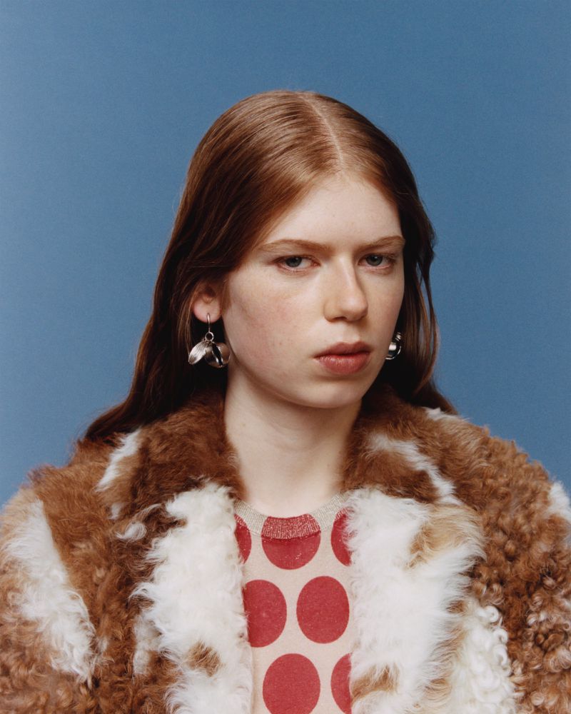 Headshot of a fair-skinned young woman with auburn hair and silver earrings, wearing a patterned top and fur coat against a blue backdrop.