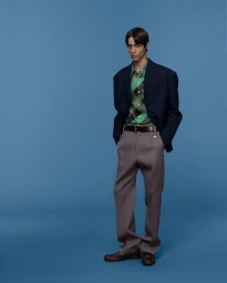 Model wearing a navy blazer, green patterned shirt, taupe trousers, and brown loafers against a solid blue studio backdrop.