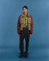 Model wearing a brown paneled bomber jacket, green collared shirt, and dark trousers against a blue seamless background.