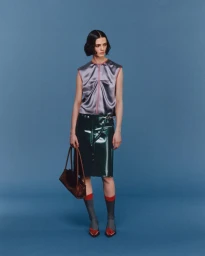 Model wearing a metallic lavender sleeveless top and glossy green skirt holding a brown leather handbag against blue seamless