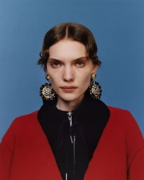 Model wearing large ornate statement earrings, a black zip top, and a red knit cardigan against a solid blue backdrop.