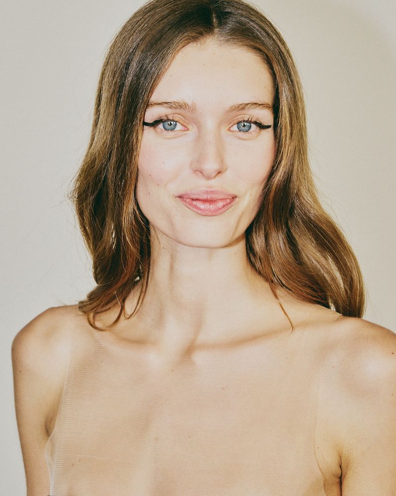 Close-up portrait of a young adult female model with light skin, long brown hair, and eyeliner, against a neutral studio backdrop.