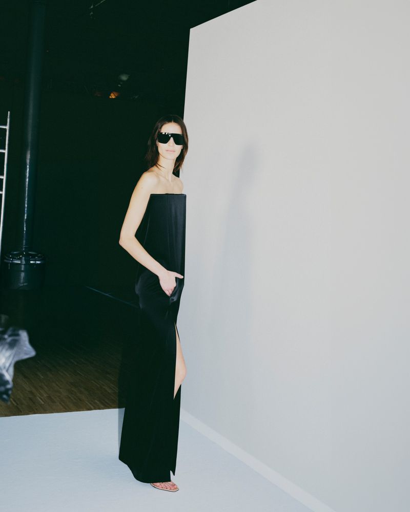 Elegant woman in a black dress and sunglasses poses against a stark white backdrop in a studio setting, showcasing minimalist fashion.