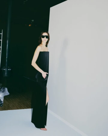 Elegant woman in a black dress and sunglasses poses against a stark white backdrop in a studio setting, showcasing minimalist fashion.