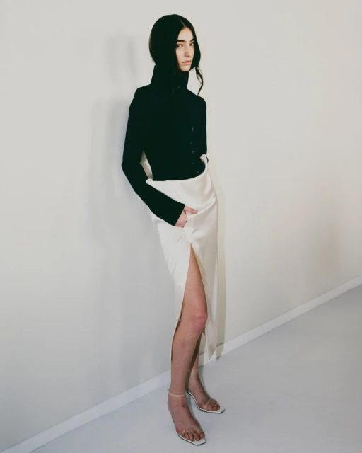 Full-length portrait of a woman in a black turtleneck and ivory skirt with a thigh-high slit, posing against a neutral wall in a studio.