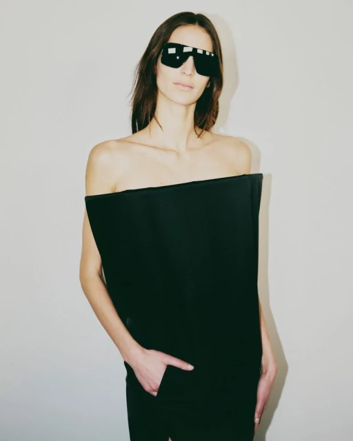 Adult female model with sunglasses in fashionable black formalwear against a plain grey backdrop. Minimalist studio portrait.