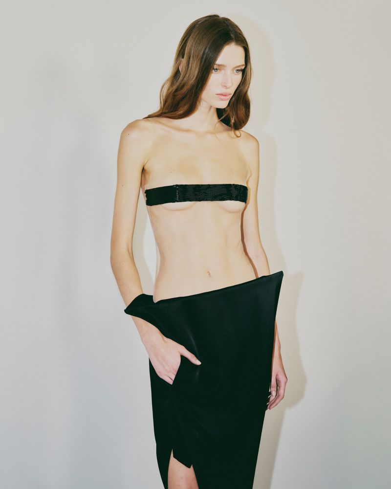 Fashion shot of a slim adult female model in an unusual black skirt with a black bandeau against a neutral studio backdrop.