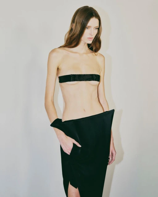 Fashion shot of a slim adult female model in an unusual black skirt with a black bandeau against a neutral studio backdrop.