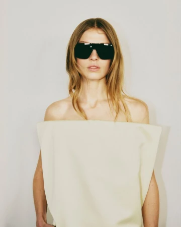 Blonde woman with sunglasses and sleeveless top poses against a white backdrop in a fashion-forward shot with neutral tones.