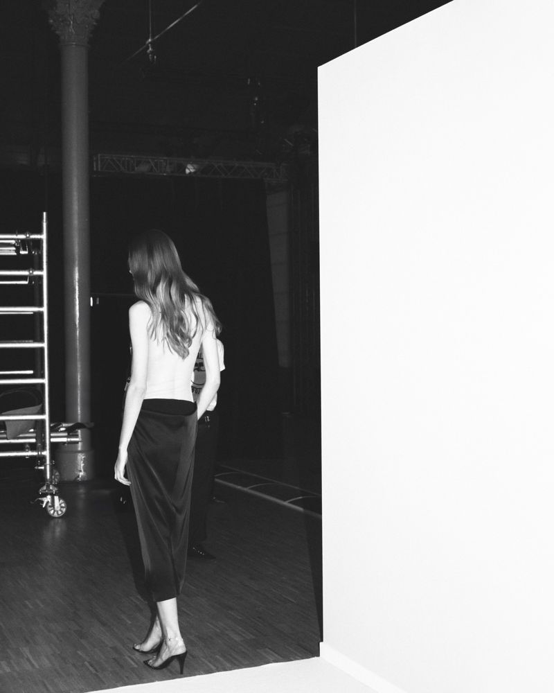 Black and white photograph of a woman in a skirt walking toward a white wall with a stage and metal scaffolding in the background.