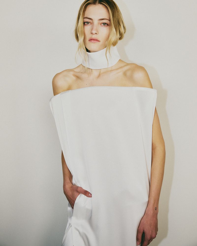 Elegant woman poses in a striking off-shoulder white dress with a high neckline, set against a clean, light backdrop in a studio shot.