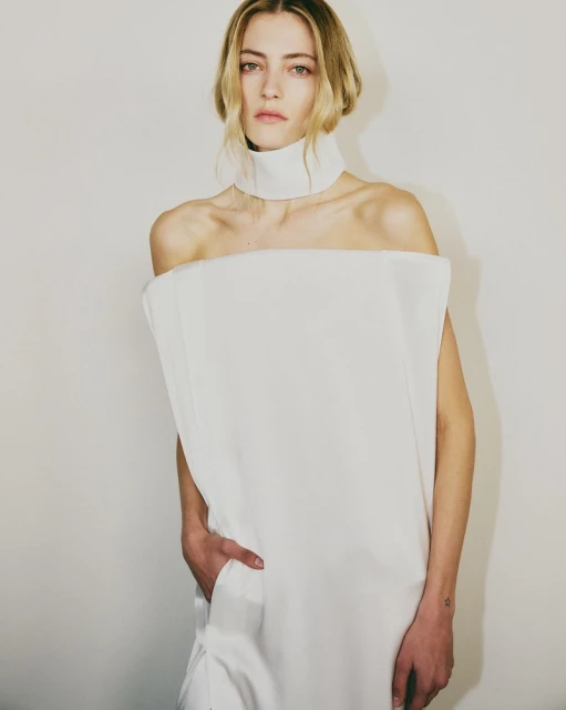 Elegant woman poses in a striking off-shoulder white dress with a high neckline, set against a clean, light backdrop in a studio shot.