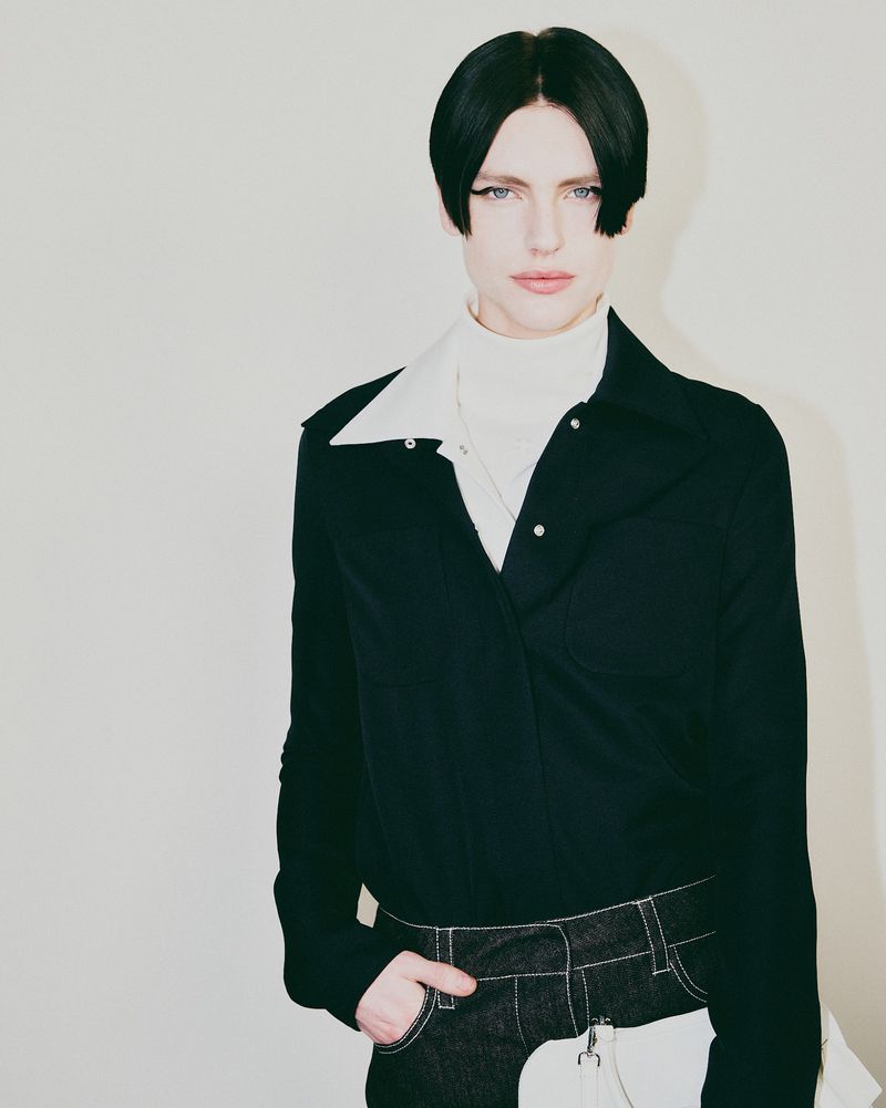 Stylish portrait of an androgynous adult with unique dark hair, wearing a black jacket over a white turtleneck, against a neutral backdrop.