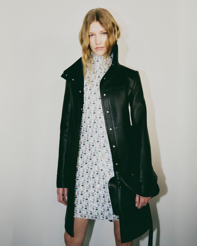 An adult female fashion model in a black coat and patterned dress poses indoors against a plain wall in a studio setting.