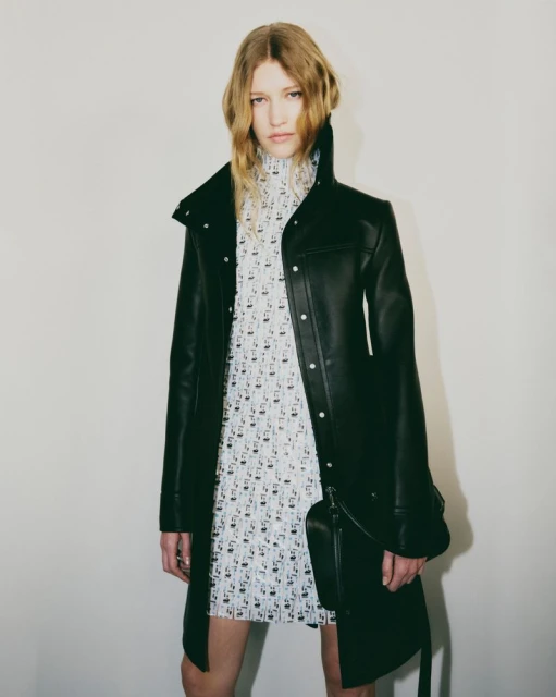 An adult female fashion model in a black coat and patterned dress poses indoors against a plain wall in a studio setting.