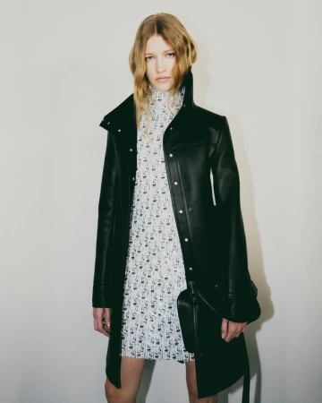 An adult female fashion model in a black coat and patterned dress poses indoors against a plain wall in a studio setting.