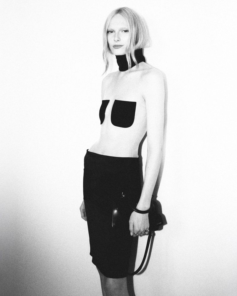 A stark black-and-white fashion portrait of a slim blonde model in a minimalist outfit, posing against a plain white background.