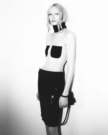 A stark black-and-white fashion portrait of a slim blonde model in a minimalist outfit, posing against a plain white background.