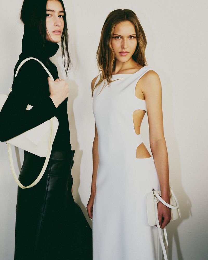 Two fashion models pose indoors against a plain white wall, showcasing a white dress and a black turtleneck outfit with matching bags.