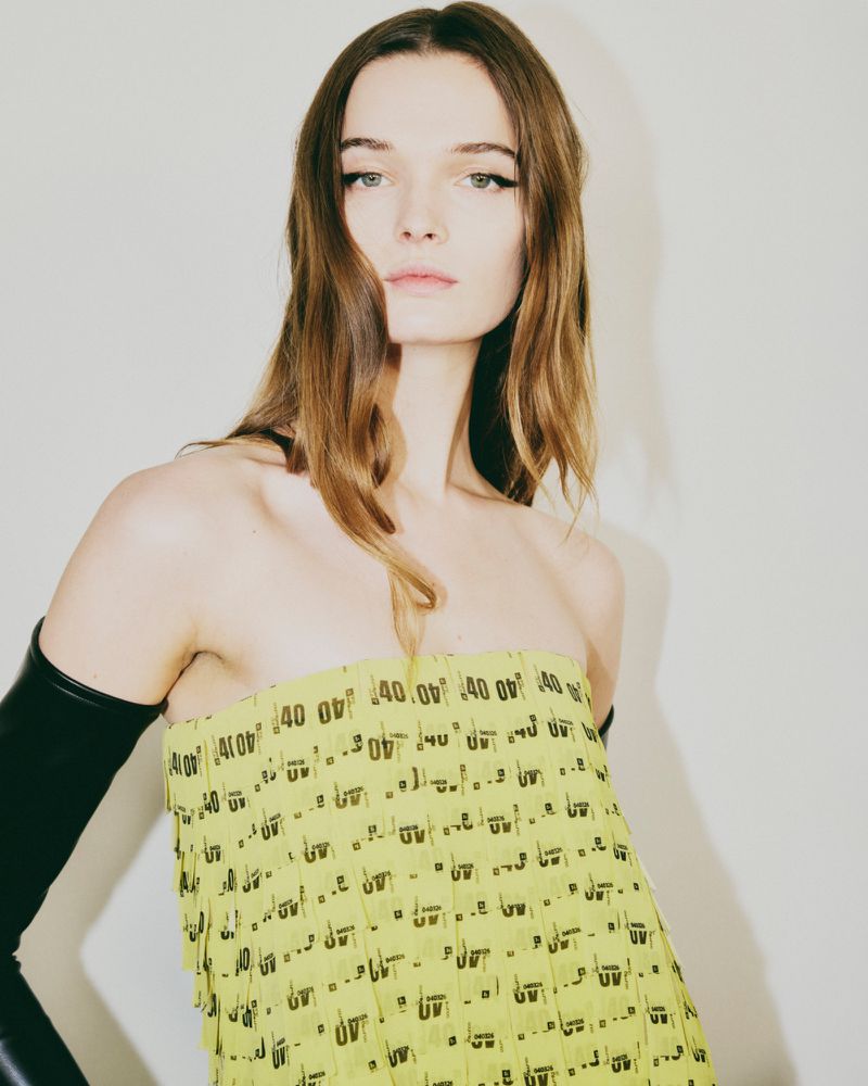 Portrait of a fair-skinned woman with long brown hair, wearing a light-green dress with black text print and black leather gloves.