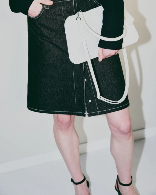 Fashion shot of woman wearing denim skirt, black long-sleeve top, black strappy heels, carrying a cream-colored handbag against a white background.
