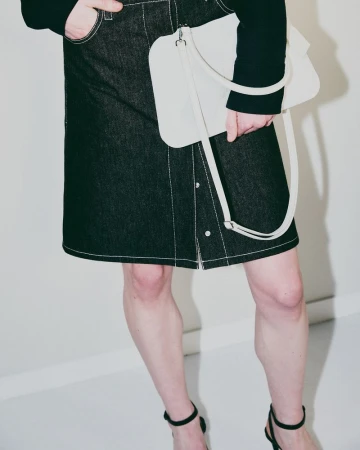 Fashion shot of woman wearing denim skirt, black long-sleeve top, black strappy heels, carrying a cream-colored handbag against a white background.