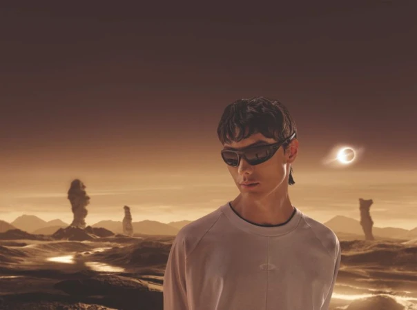 A model with sunglasses poses in a long-sleeve shirt, set against an out-of-focus desert landscape with rock formations and an eclipse.