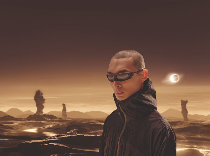 Asian man in sunglasses and hooded jacket stands in a desert landscape with rock formations and a partial solar eclipse in the background.