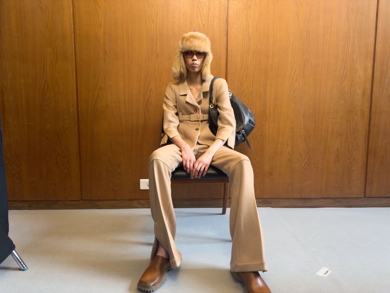 A woman in a beige suit and fur hat sits indoors. Her look is complemented by brown shoes and a black backpack, set against a wooden wall.