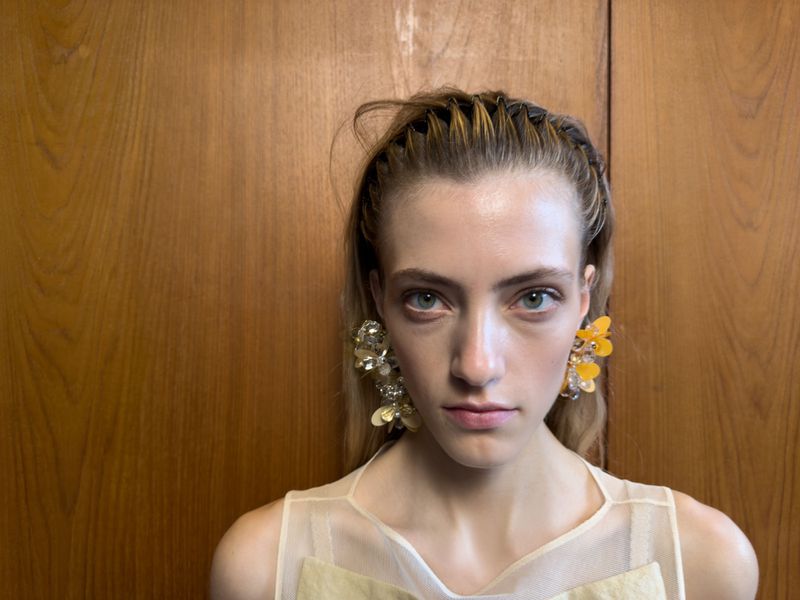 Close-up portrait of a young woman with braided hair, wearing statement earrings, and a light-colored sleeveless top against a wooden background.