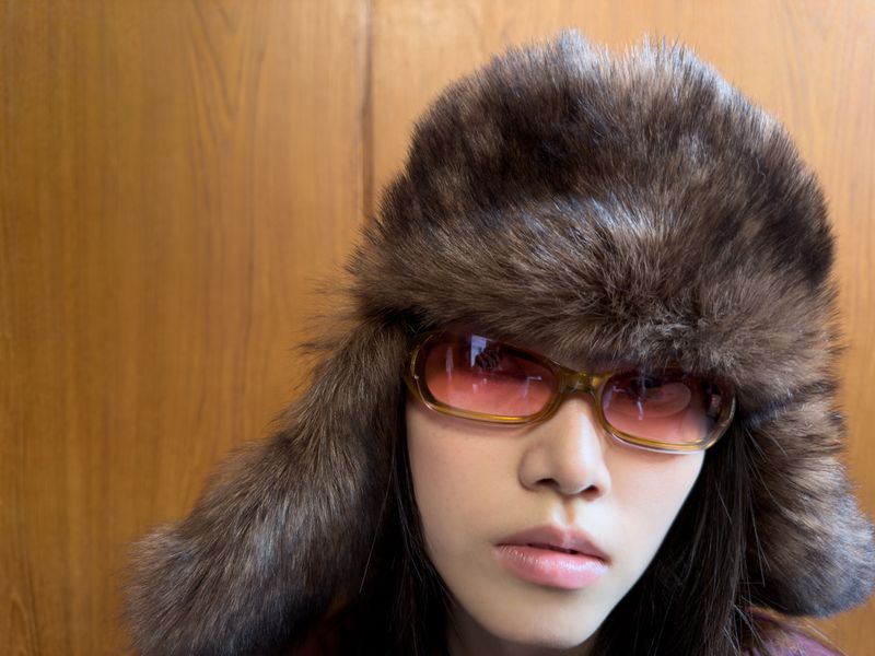 Woman with sunglasses and a fur hat poses indoors against a wooden panel. Fashion shot with warm colors and a neutral facial expression.