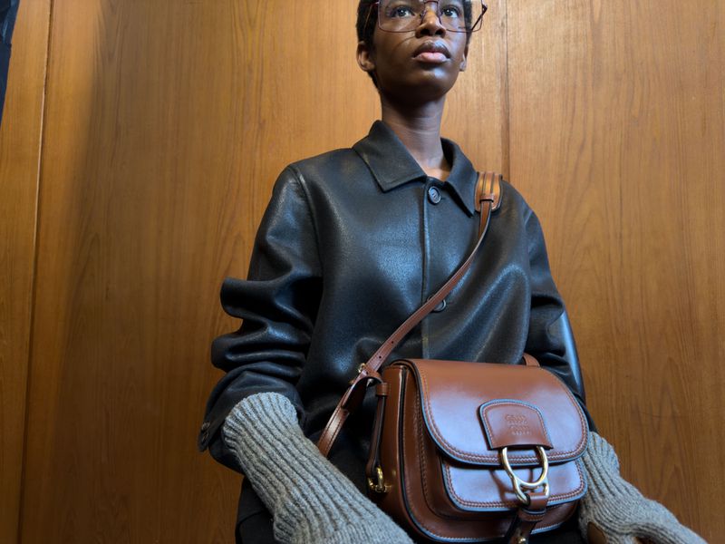 Stylish woman in leather jacket and tan purse poses indoors against a wood paneled wall in a fashion forward editorial portrait.