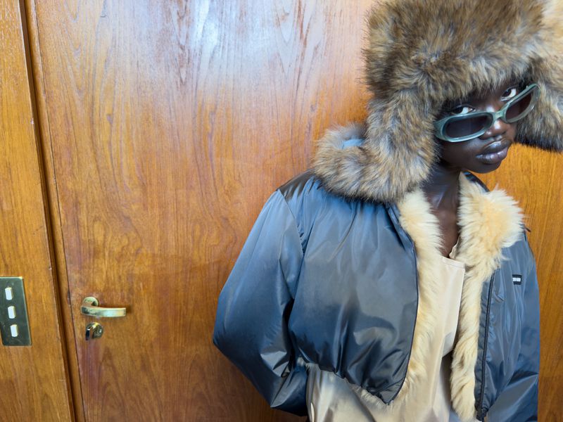 A person models a fur-lined hat and jacket in front of a wooden door, showcasing a fashion-forward style and cool color palette.