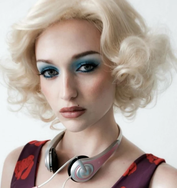 Portrait of a young woman with blonde curly hair and blue eye shadow wearing a floral dress and headphones in a white background.