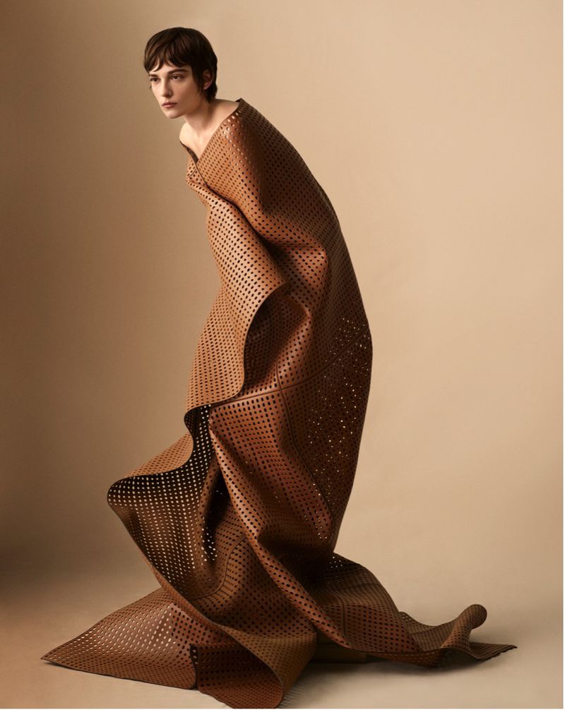 Fashion shot of a woman draped in a brown, perforated fabric, posing in a studio. The neutral backdrop accentuates the textured material.