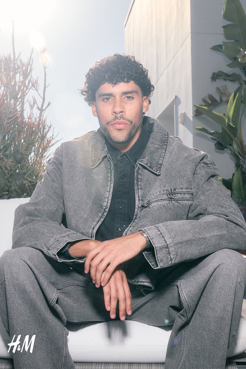 Man poses outdoors wearing grey denim jacket and pants, seated on a white bench with plants and a building visible in the background.