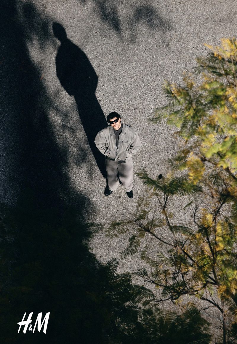 Aerial view of a man in a grey jumpsuit standing on a road with trees casting shadows and the H&M logo visible.