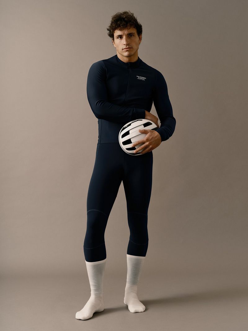Male cyclist in form-fitting navy blue athletic wear holds a white and black helmet in a studio shot on a neutral background.