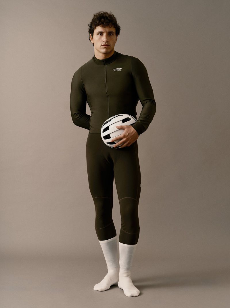 Male cyclist in olive green jumpsuit holds white helmet in a studio shot against a neutral background with natural light.