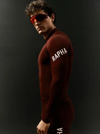 Male cyclist posing in cycling gear and sunglasses against a dark gray background in a studio setting, showcasing sportswear fashion.