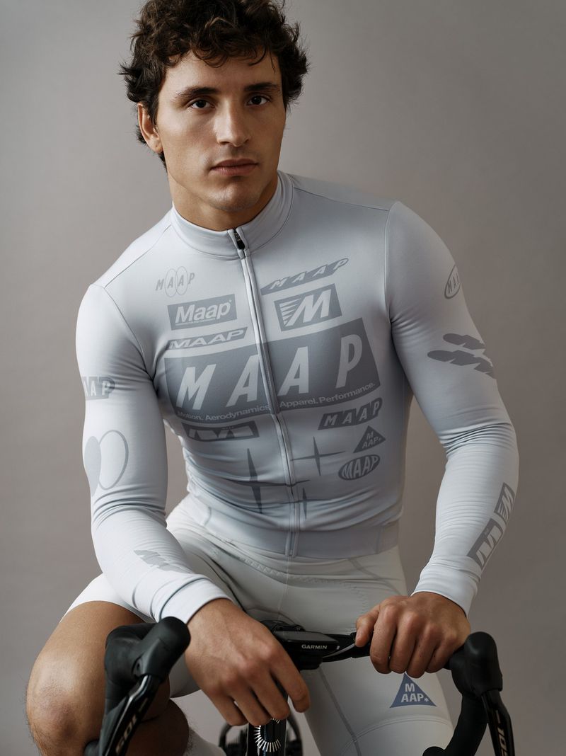 Athletic man in a light grey cycling jersey poses seated on a bicycle with hands on handlebars, against a solid grey backdrop.