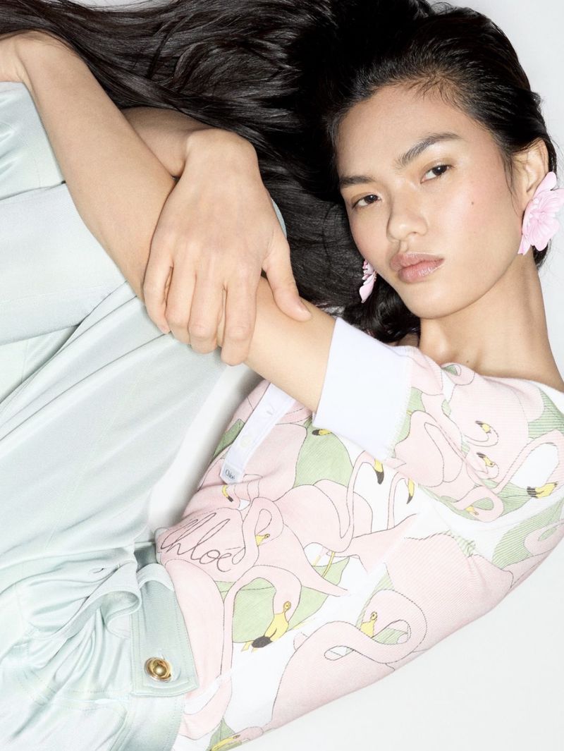 Woman with long black hair lying down wearing a flamingo pattern top and pale green skirt. Flower earrings, indoor setting, eye-level view.