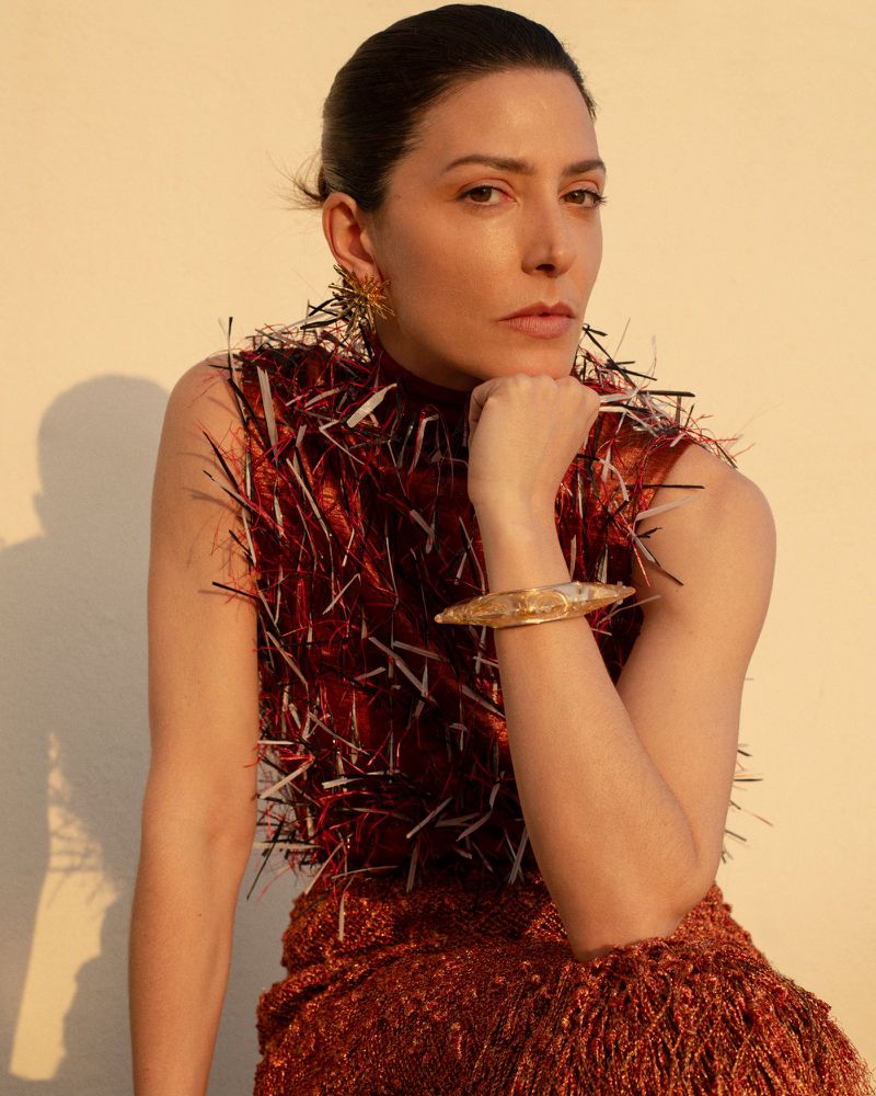 Portrait of a woman in a red fringed dress and gold jewelry, looking at the camera in soft lighting with a cream-colored background.