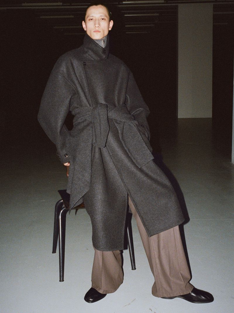 A person poses in a grey wool coat with wide-legged brown trousers and black shoes, standing next to a chair, in a dimly lit indoor setting.
