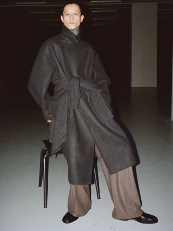 A person poses in a grey wool coat with wide-legged brown trousers and black shoes, standing next to a chair, in a dimly lit indoor setting.
