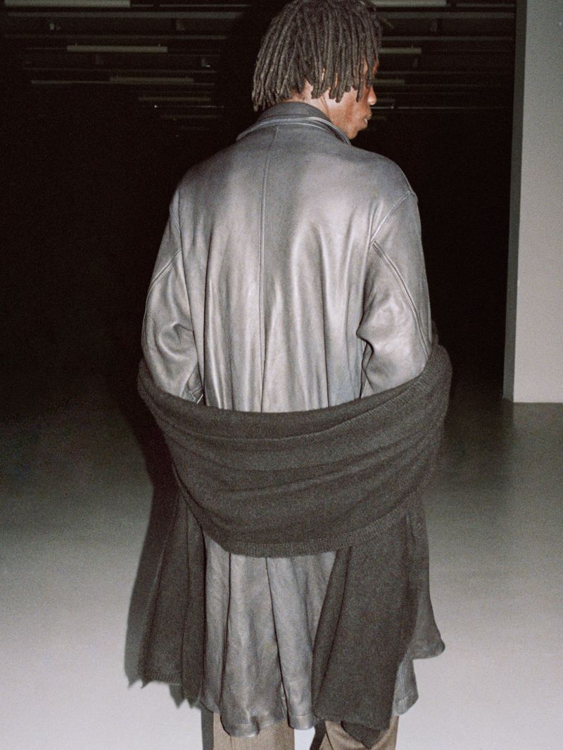Back view of an adult male with dreadlocks wearing a long grey leather coat and black scarf indoors against a dark grey and white background.