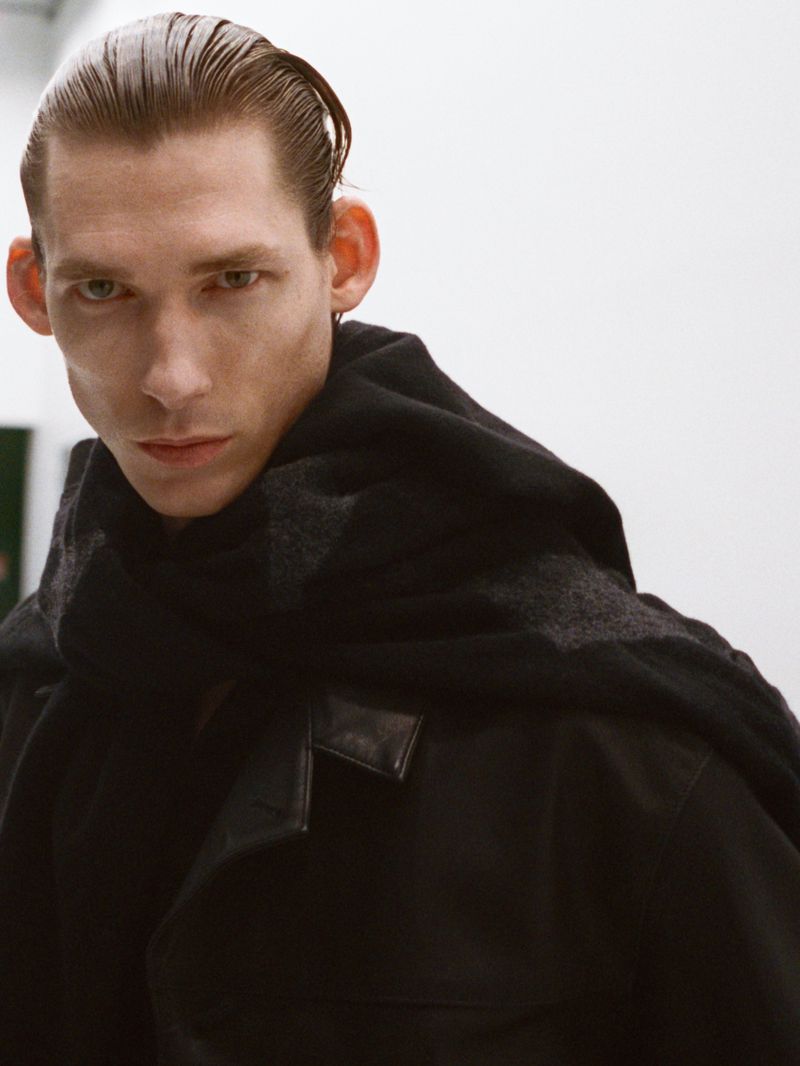 Male model with slicked-back hair wearing an all-black jacket and heavy wrap against a plain background.